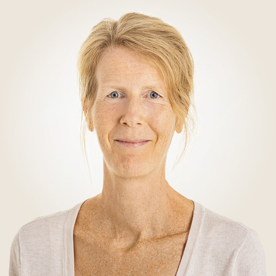 Eine lächelnde Frau mit blauen Augen und hochgesteckten blonden Haaren, die Natürlichkeit und Authentizität ausstrahlt.