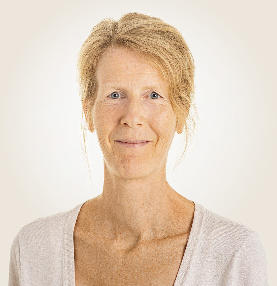 Eine lächelnde Frau mit blauen Augen und hochgesteckten blonden Haaren, die Natürlichkeit und Authentizität ausstrahlt.